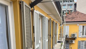 Un balcone stretto con persiane chiuse, ringhiera in metallo e vista sui palazzi vicini.