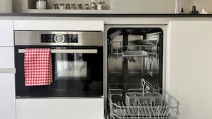 Una cucina con forno Bosch e lavastoviglie, un asciugamano a quadretti bianchi e rossi appeso alla porta del forno e un portascopetti metallico all'interno della lavastoviglie.
