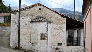 Ein kleines, rustikales Gebäude mit einem Ziegeldach und einem Kreuz darauf, wahrscheinlich eine Kapelle oder eine kleine Kirche, mit einem Schild, das es als 'The Teacher's House' kennzeichnet.