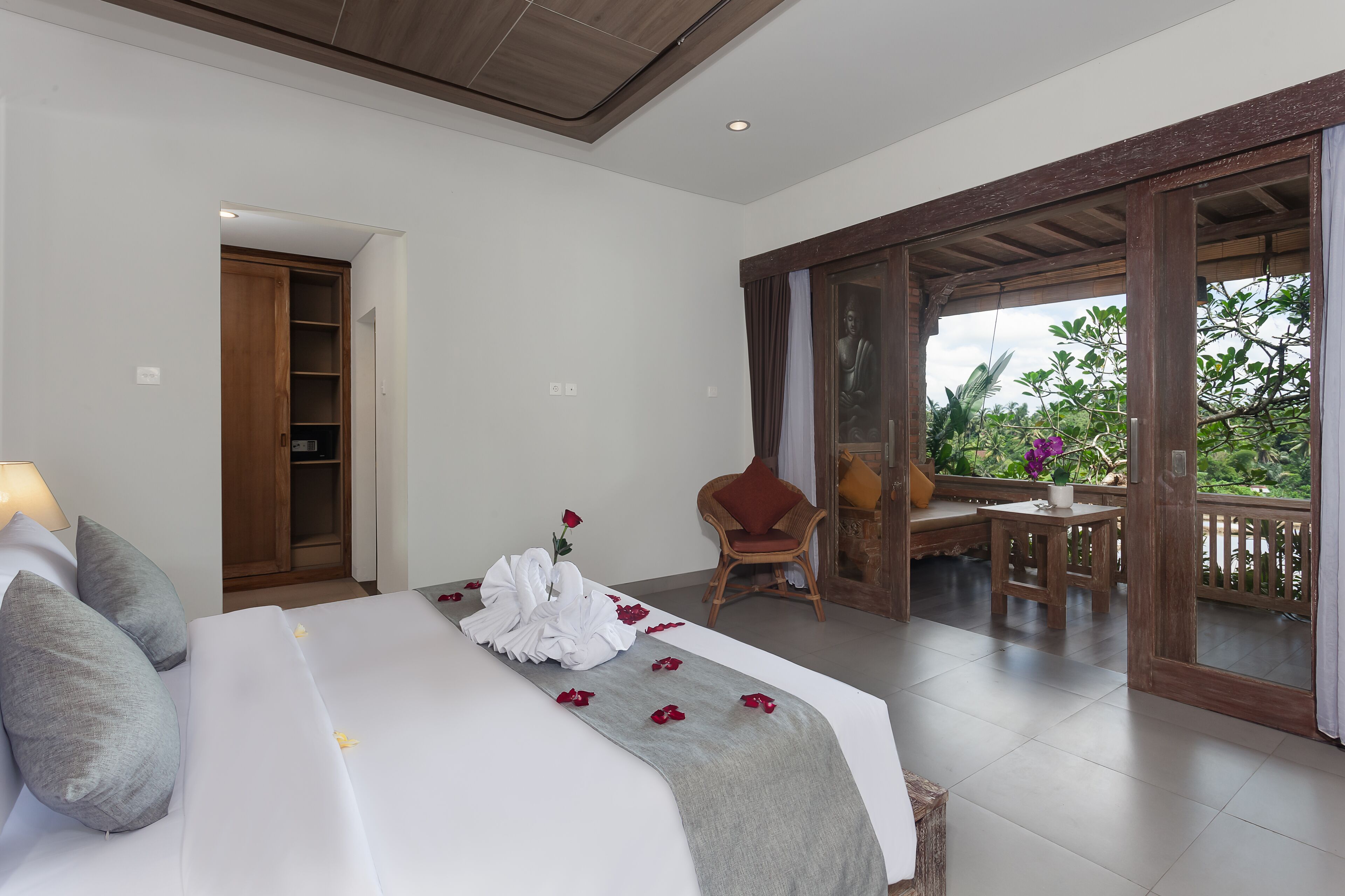 A hotel room with a bed, a chair, a table, and a balcony with a view of greenery.