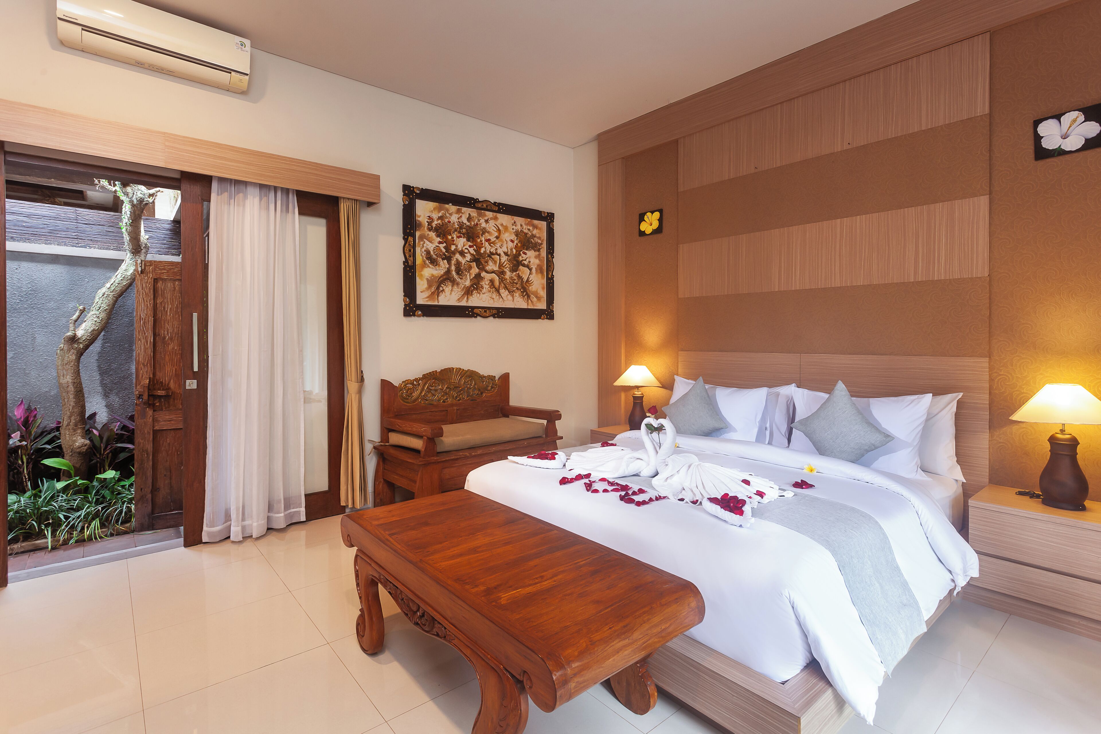 A hotel room with a bed, bedside tables, a wooden bench, a chair, a wall-mounted air conditioner, a framed artwork, and a door with a view of plants.