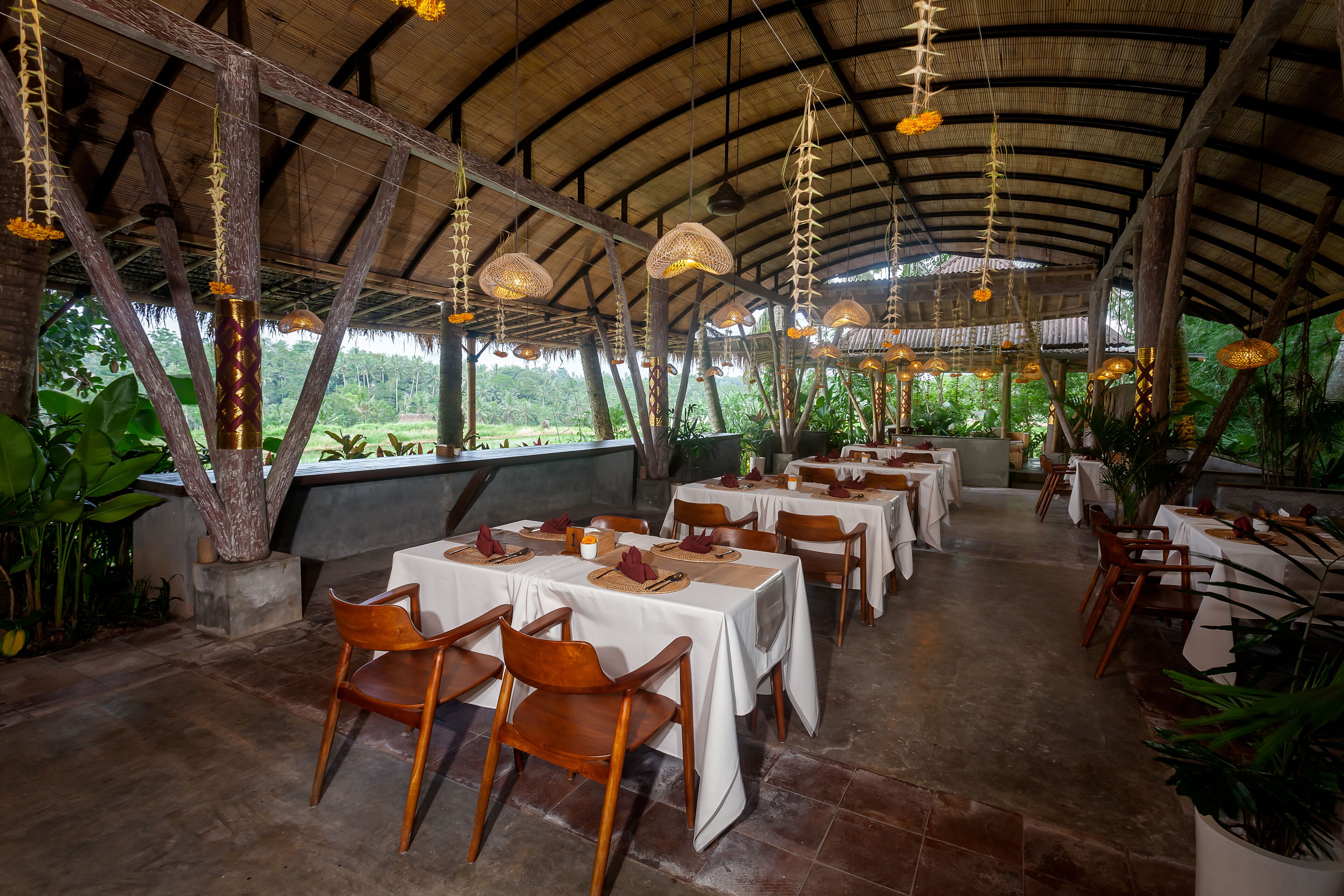 An indoor dining area with tables and chairs, a high ceiling with hanging lights, and a view of greenery outside.