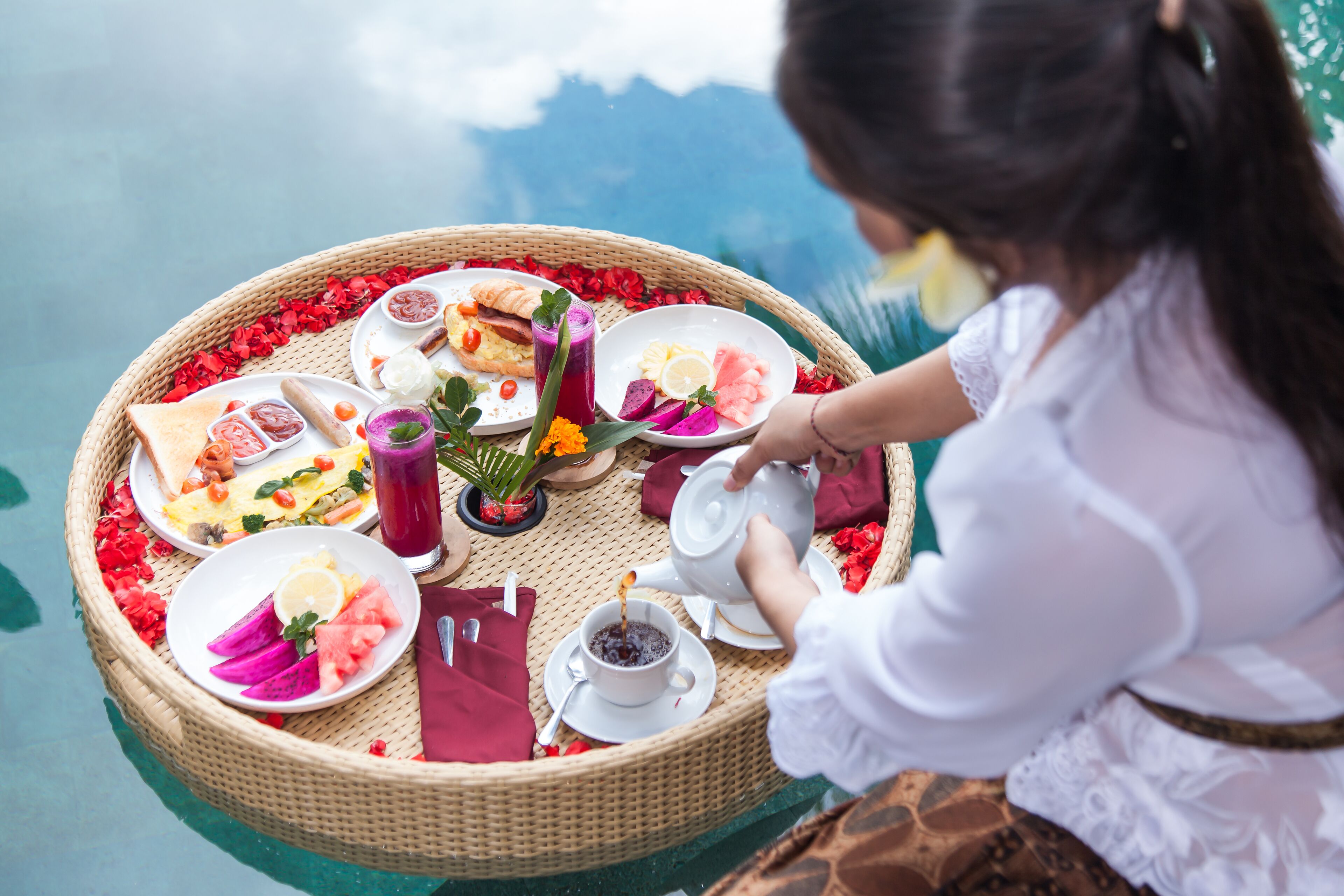 A floating dining setup with a variety of dishes and beverages, including a teapot and cups, surrounded by red flowers.