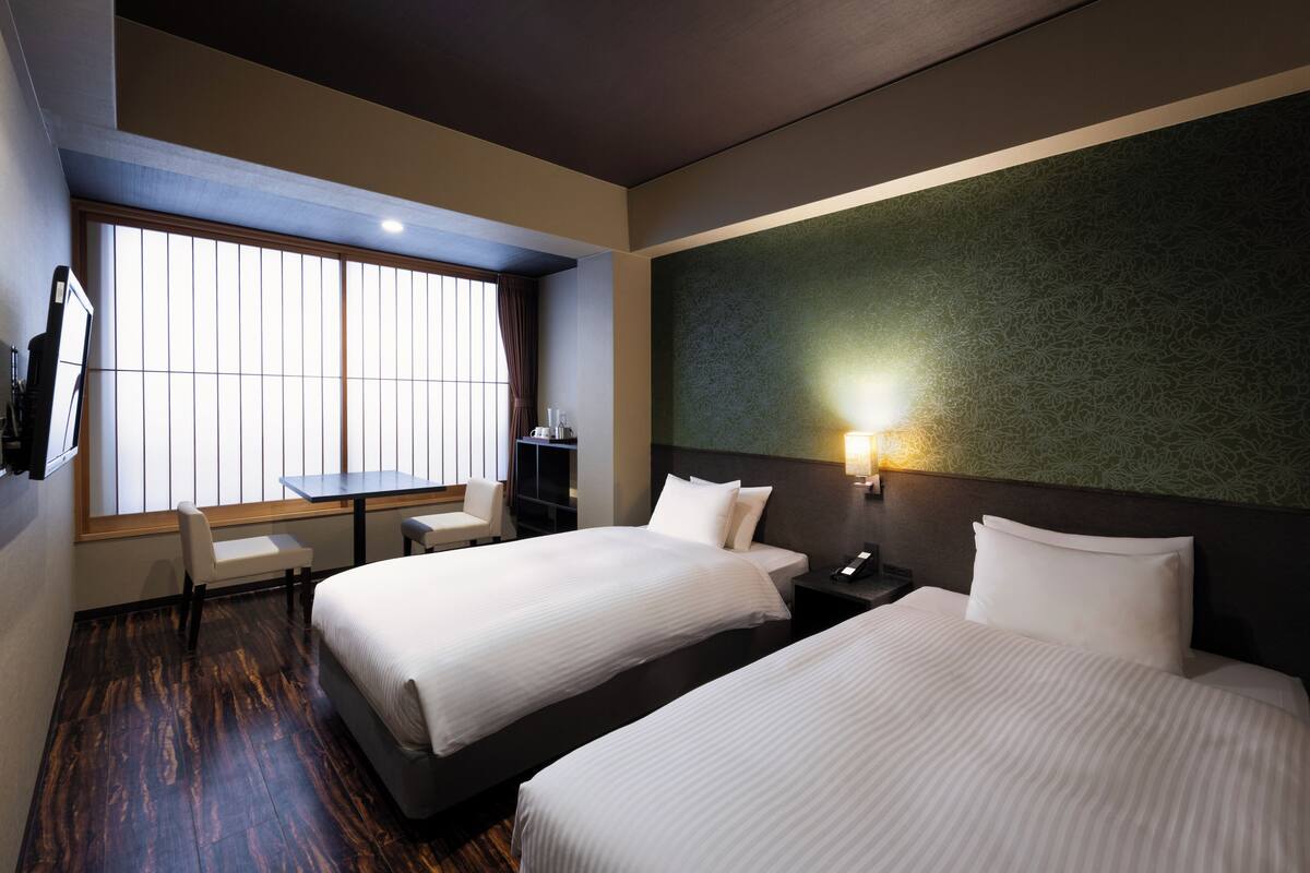 A hotel room with two beds, a desk, a chair, and a large window.