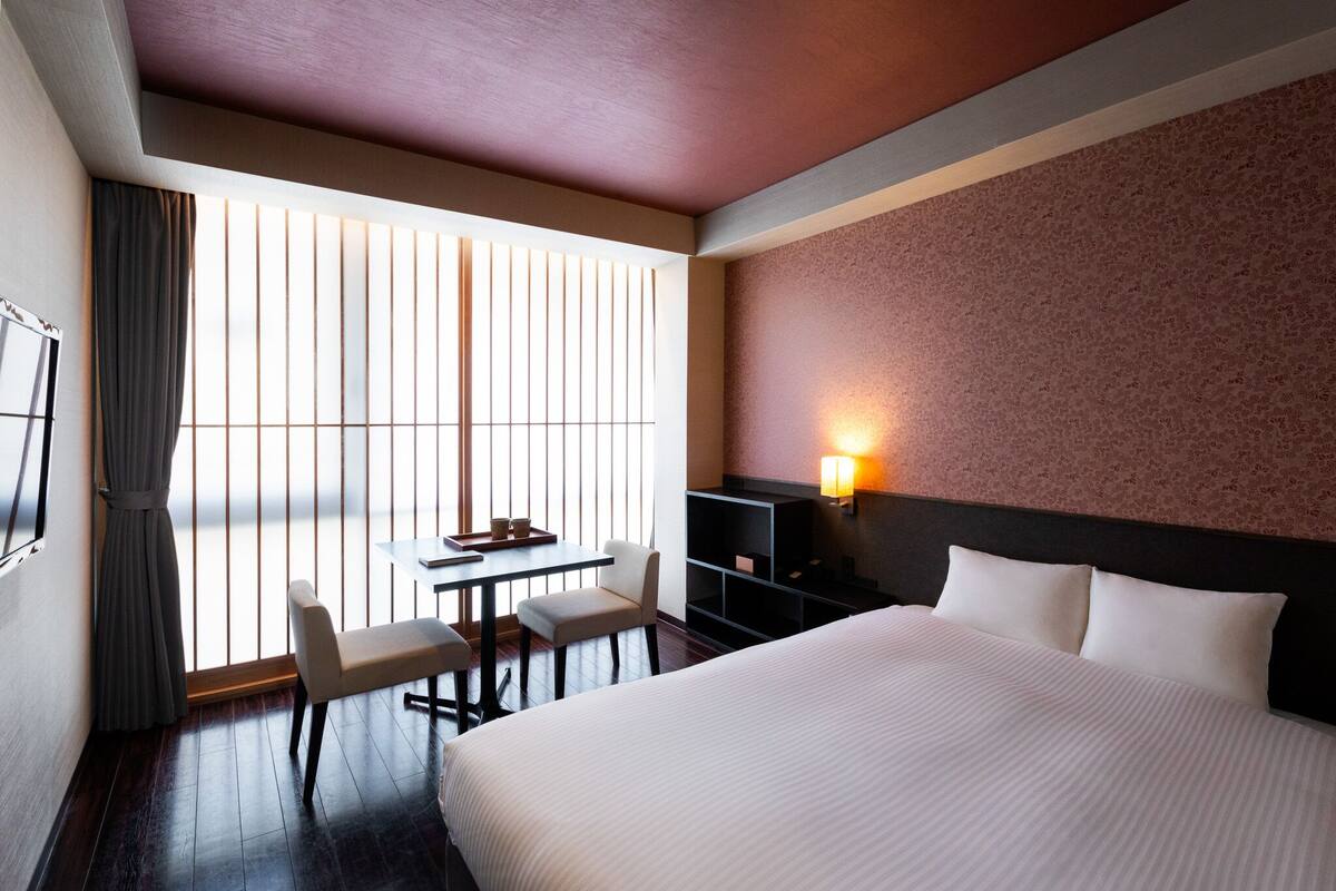 A hotel room with a large bed, a desk with chairs, a TV, and a window with blinds.