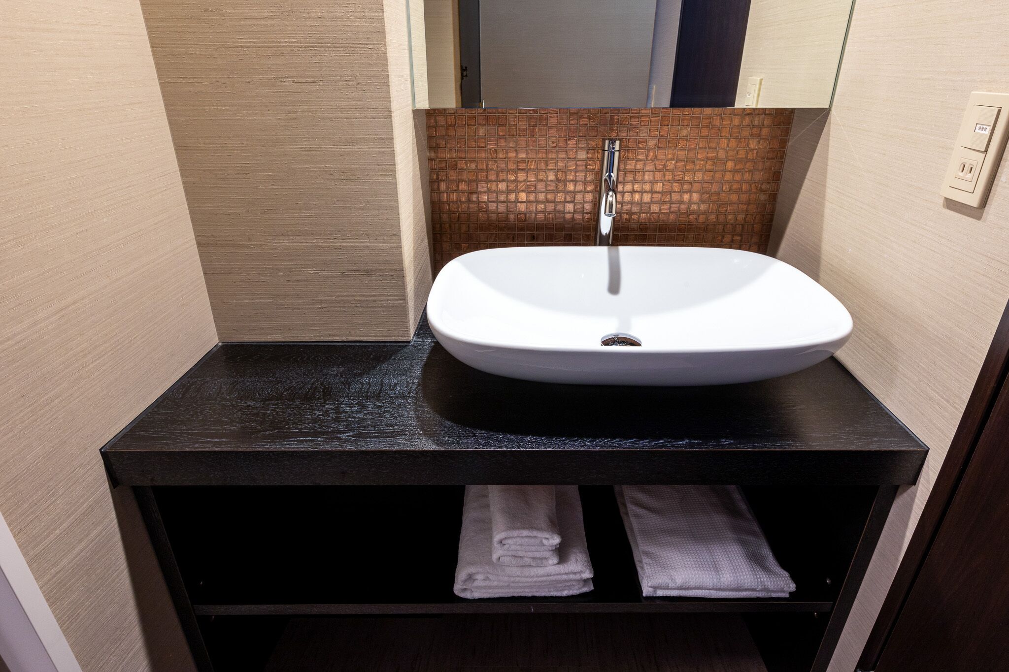 A bathroom with a white sink, black countertop, and a mirror above it. There are two towels in a drawer below the sink.