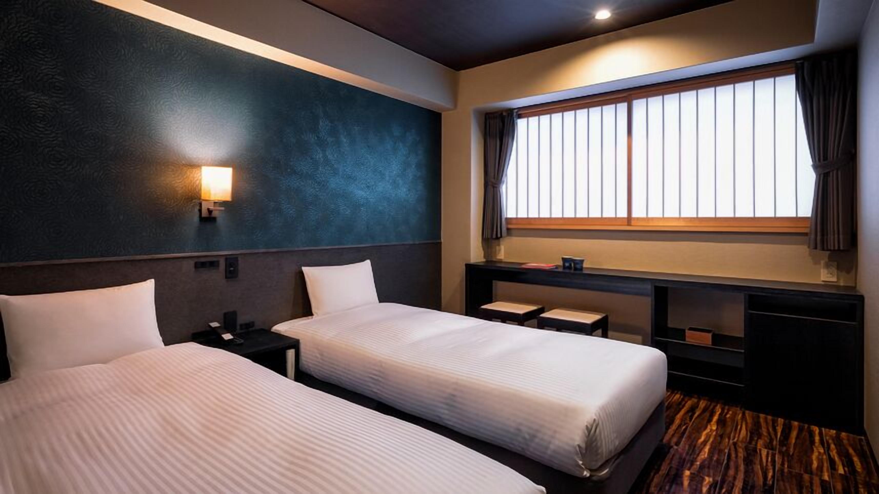 A hotel room with two beds, a nightstand, a desk, a window with curtains, and a wall-mounted lamp.