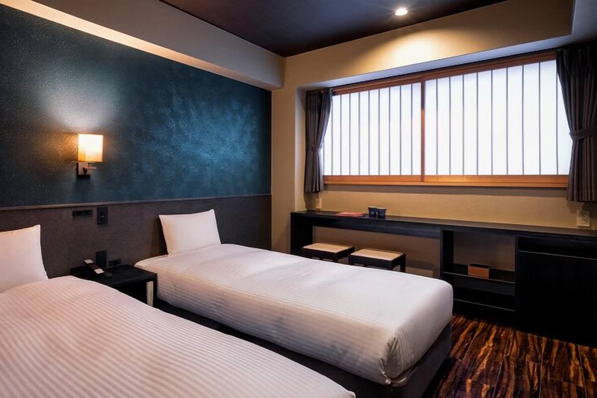 A hotel room with two beds, a nightstand, a desk, a window with curtains, and a wall-mounted lamp.