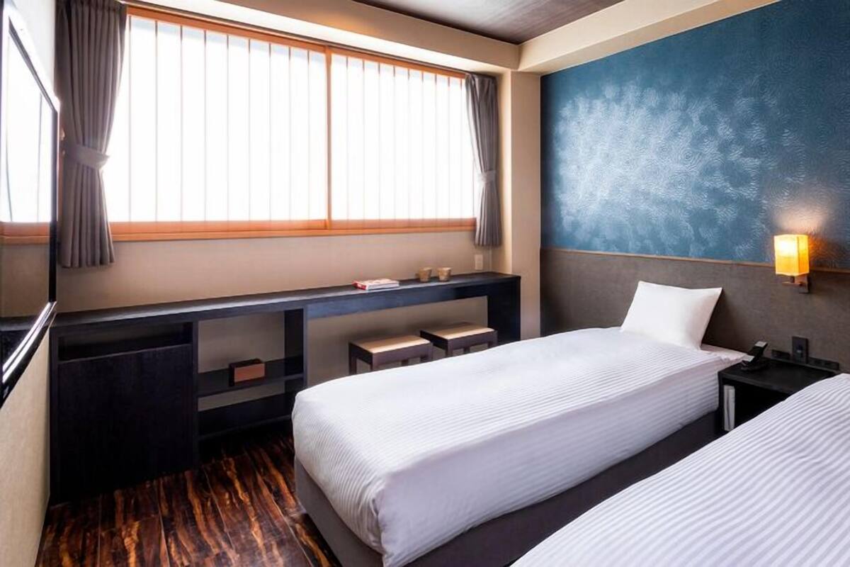 A hotel room with two beds, a desk, a chair, a nightstand, and a window with curtains.