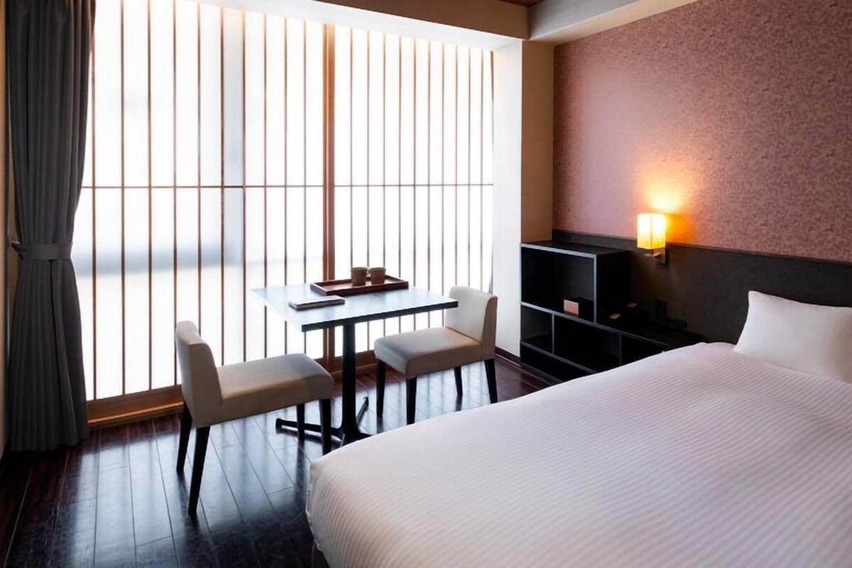 A hotel room with a bed, a desk with two chairs, a small table, a TV stand, and a large window with blinds.