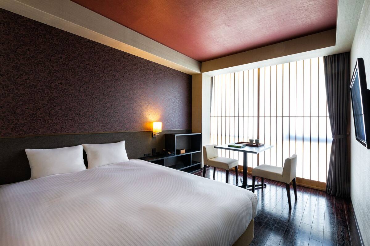 A hotel room with a large bed, a desk with a chair, a TV, and a window with blinds.