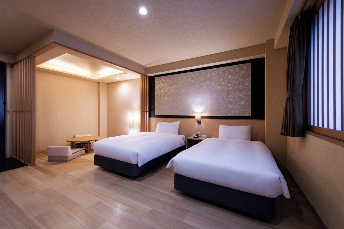 A hotel room with two beds, a small table, a chair, a large artwork on the wall, and a window with curtains.