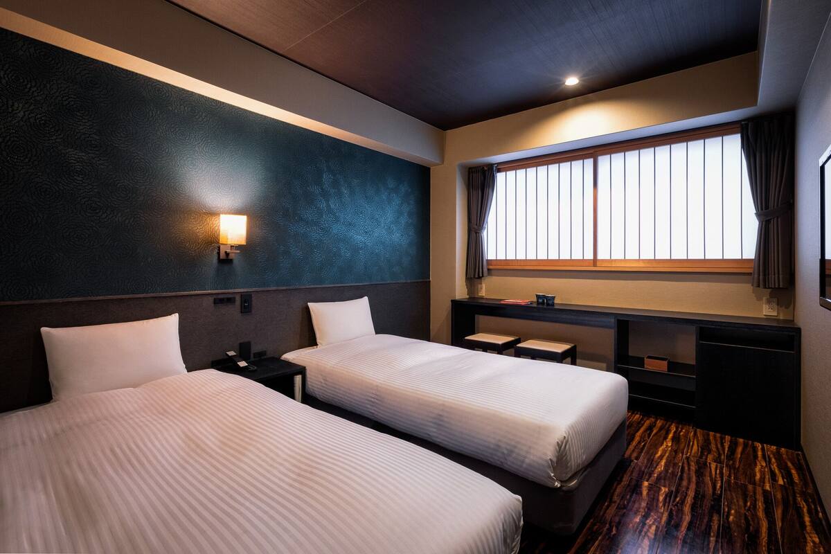 A hotel room with two beds, a nightstand, a desk, a window with curtains, and a wall-mounted light.