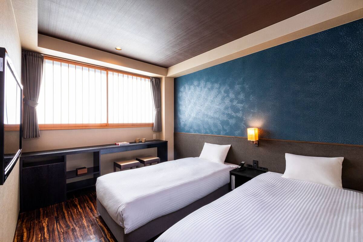 A hotel room with two beds, a desk, and a large window with curtains.