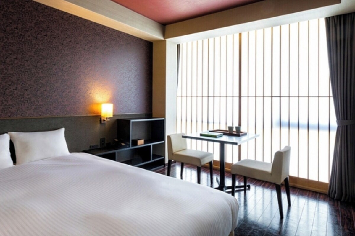 Une chambre d’hôtel avec un grand lit, un bureau avec deux chaises, une télévision et une fenêtre avec des stores verticaux.