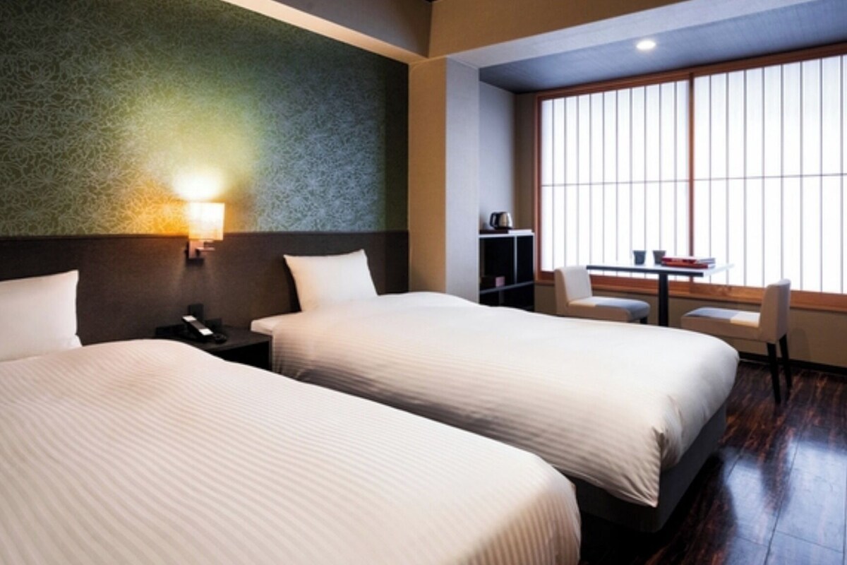 Une chambre d’hôtel avec deux lits, un bureau, une chaise et une fenêtre avec des stores.