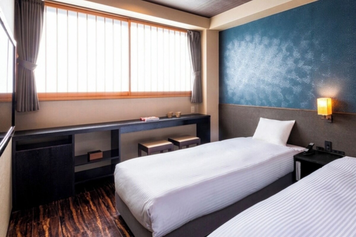 Une chambre d’hôtel avec deux lits, un bureau avec une chaise, une fenêtre avec des rideaux et une lampe fixée au mur.