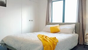 Una cama bien tendida con ropa de cama blanca y amarilla, un cabecero gris y una ventana con cortinas.