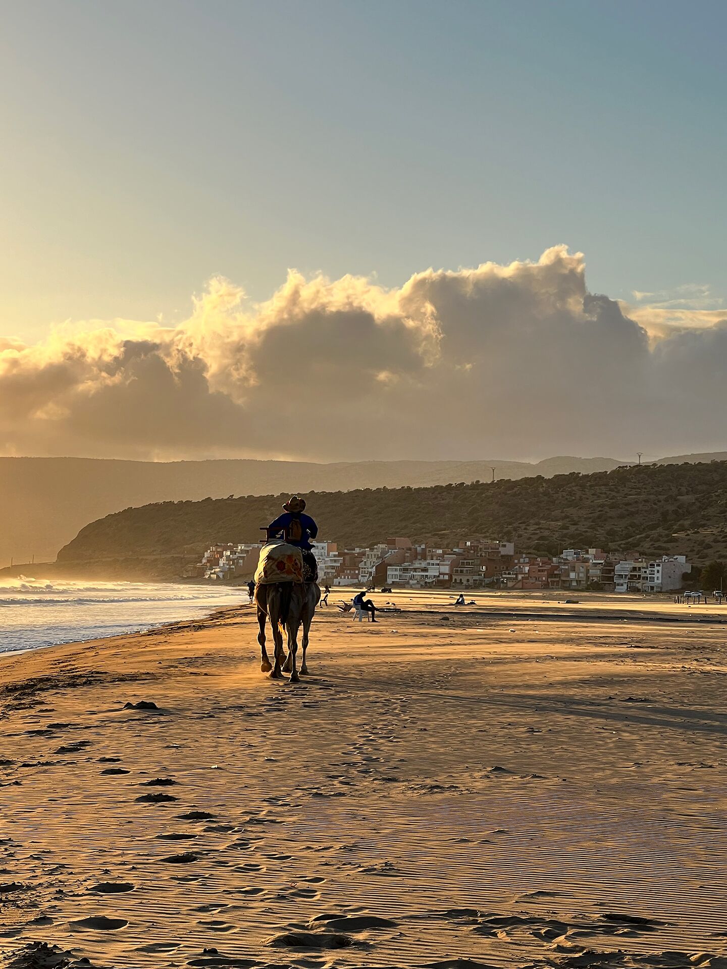 Cena de praia com uma pessoa cavalgando um camelo, um caminho de areia e edifícios ao fundo.
