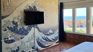 Ett hotellrum med en traditionell japansk målning av vågor på väggen, en platt-TV monterad ovanför sängen och utsikt över havet genom stora fönster.