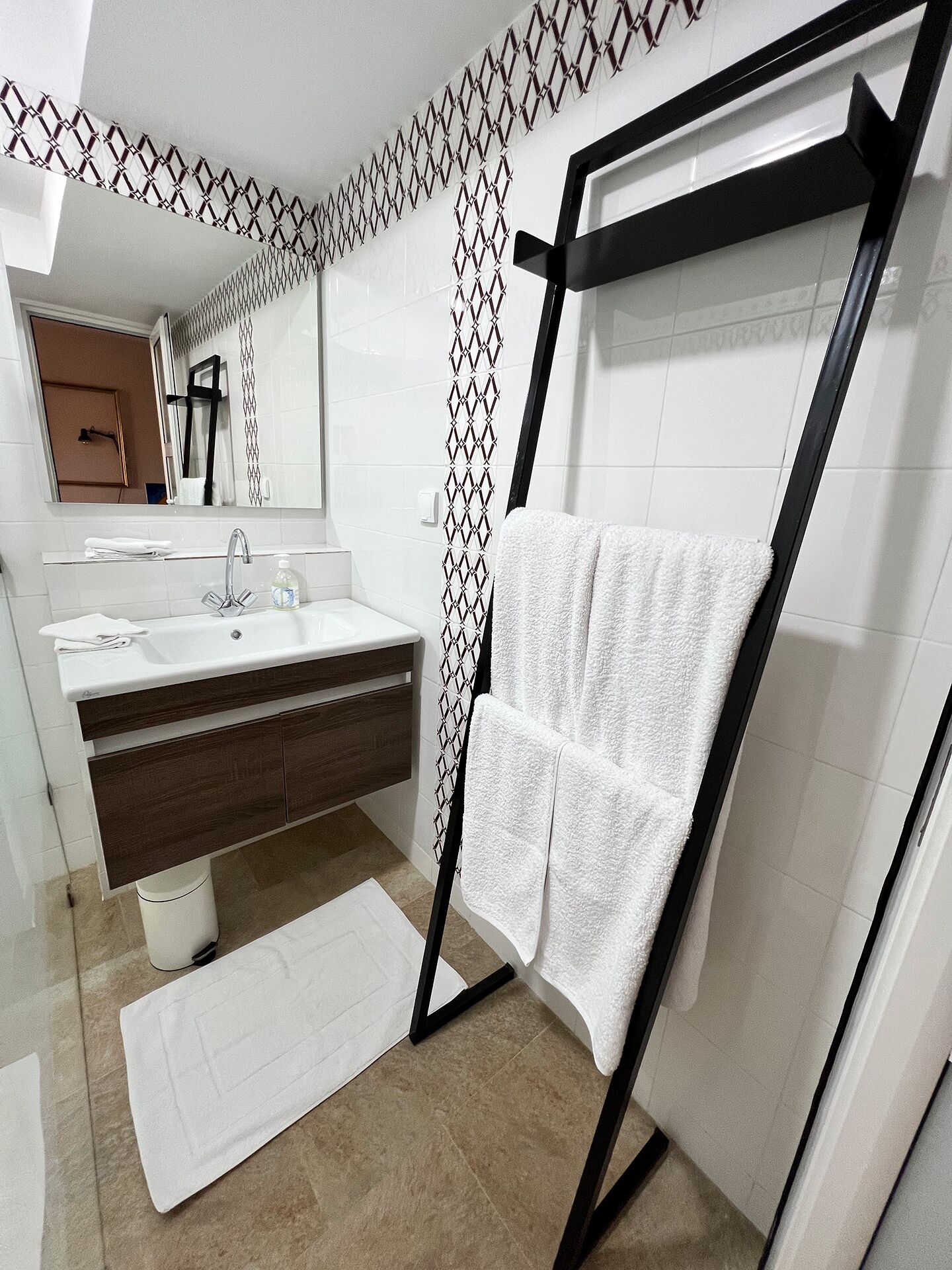 Un baño moderno con un lavamanos blanco, tocador de madera oscura, un perchero negro y azulejos con diseño.