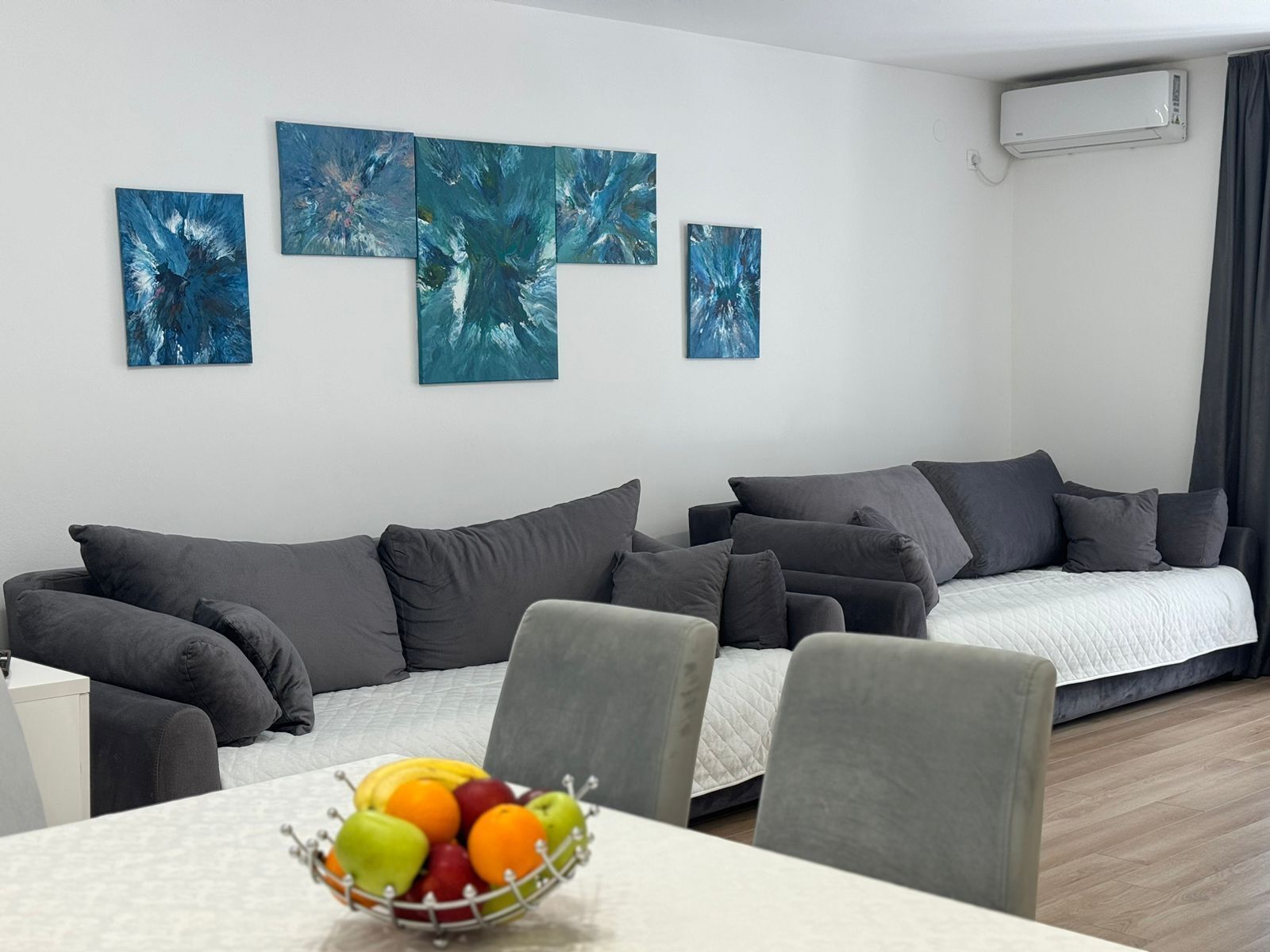Una sala de estar moderna con un sofá gris, una mesa de comedor con frutero, y arte abstracto en la pared.