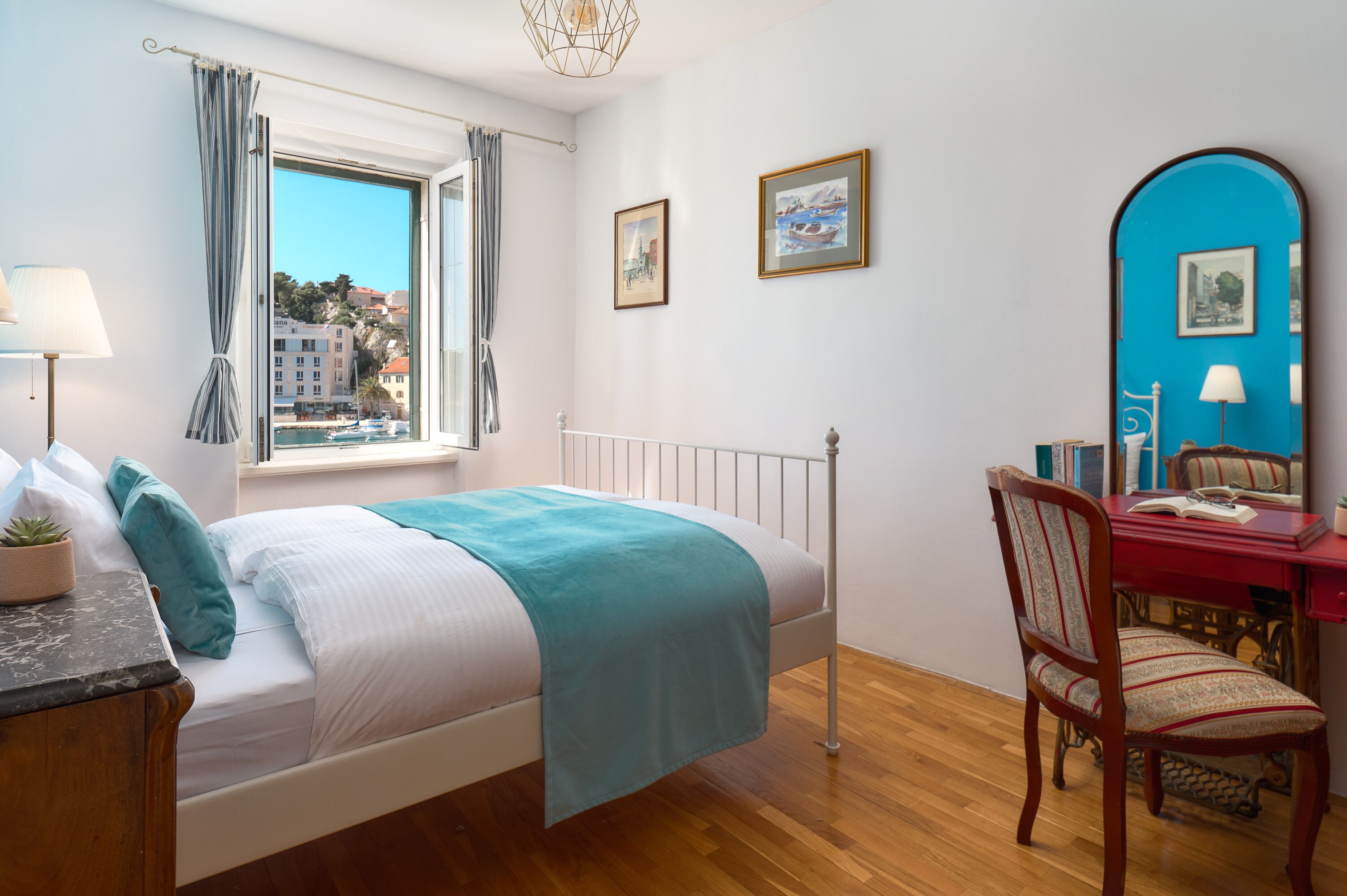Ein Schlafzimmer mit Bett, Schreibtisch, Stuhl, einem Fenster mit Vorhängen und Blick auf Gebäude.