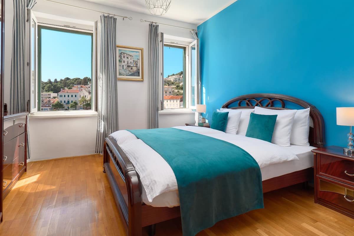 A bedroom with a large bed, a view of buildings through a window, and a blue wall.