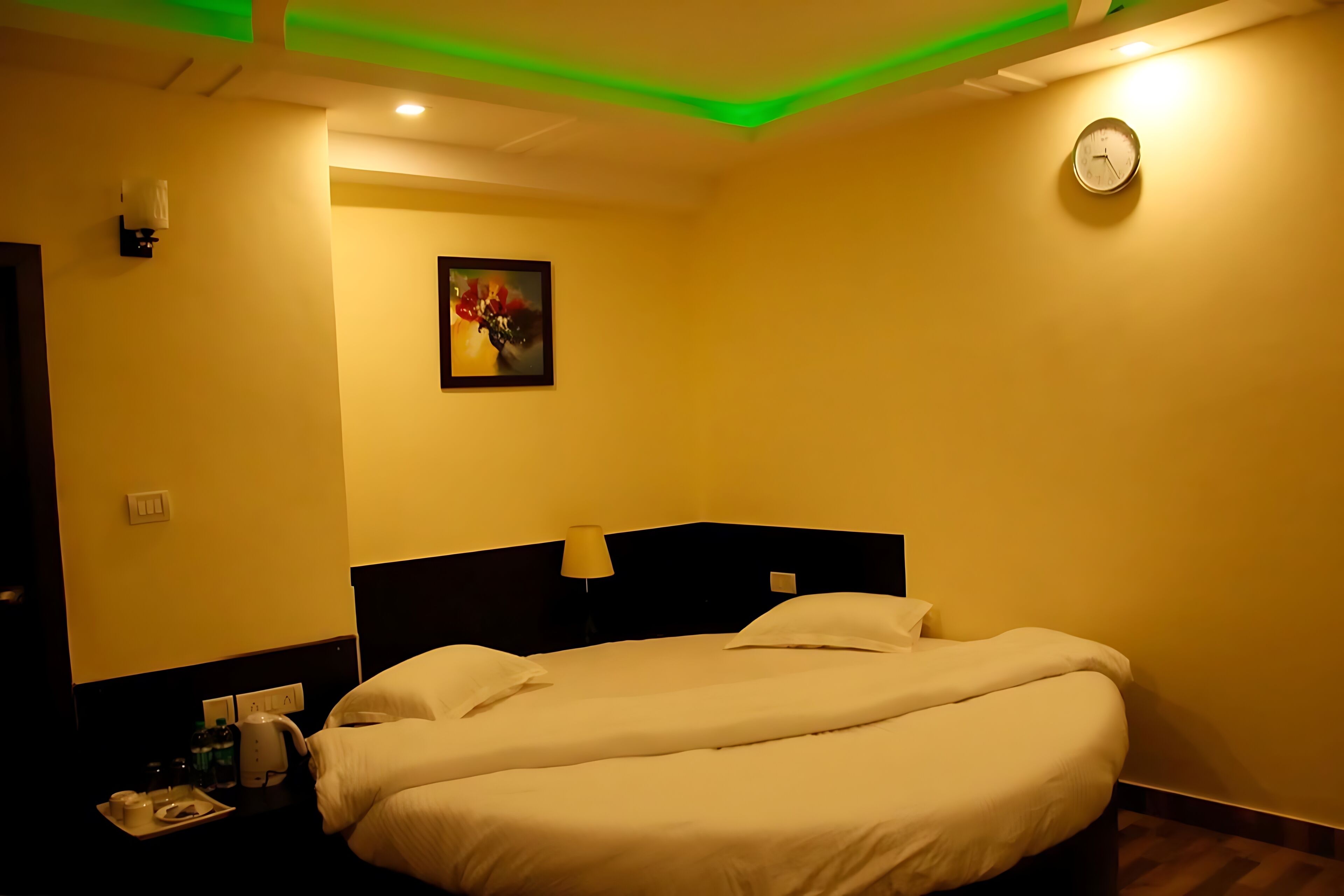 A hotel room with a bed, bedside table, lamp, clock, and a painting on the wall.