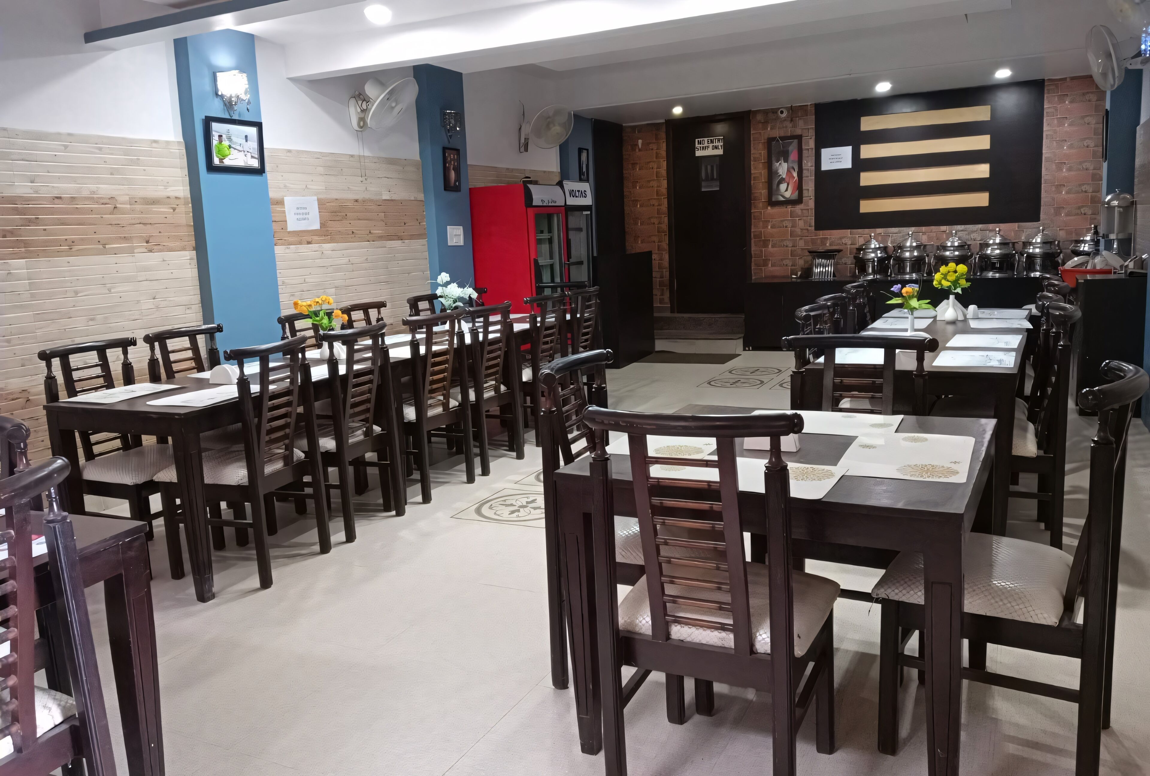 A restaurant with multiple wooden tables and chairs, a kitchen area with pots and pans, and a blue and brick wall design.