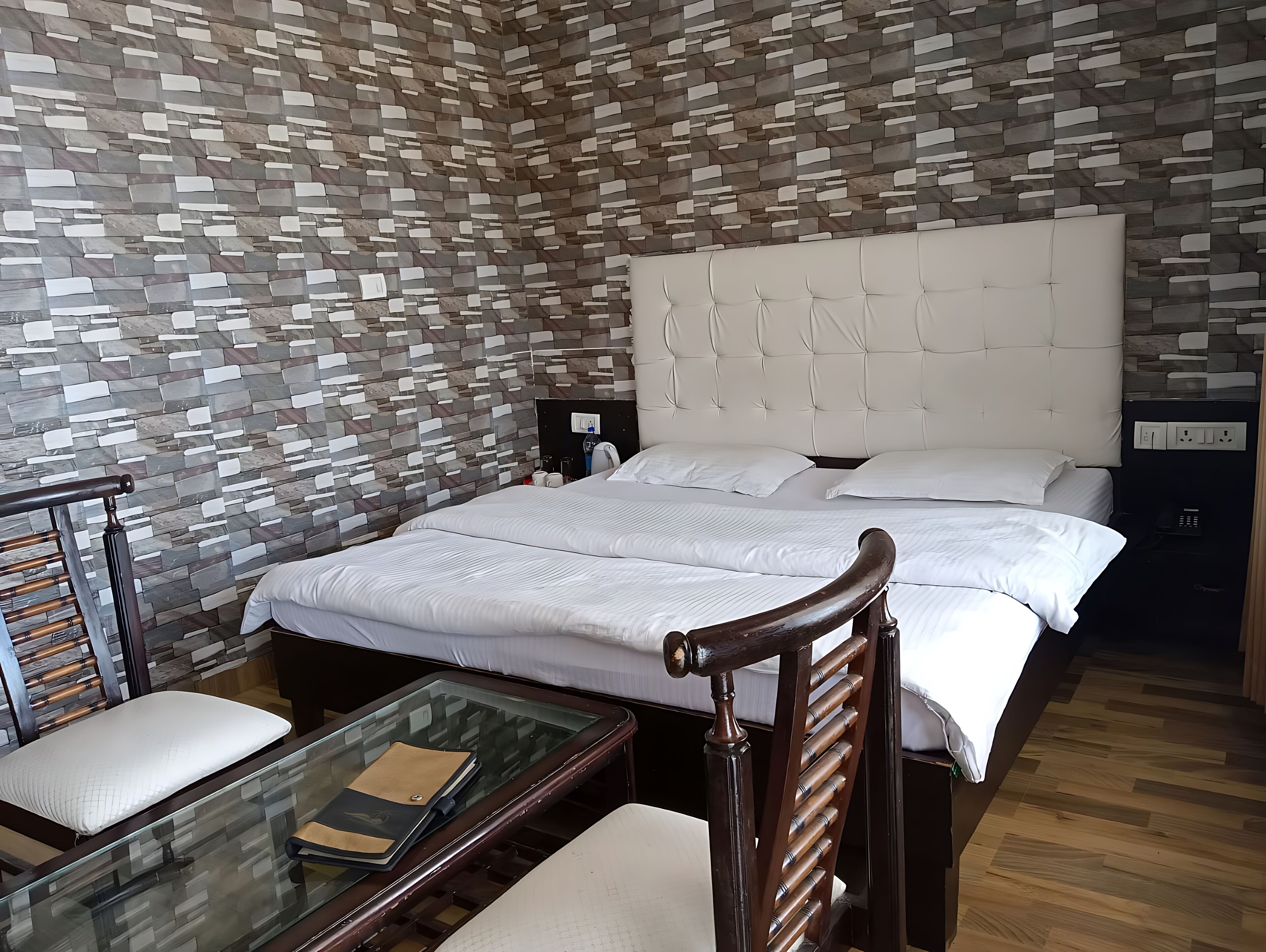 A modern hotel room with a stone-patterned wall, a tufted headboard, a glass coffee table, and a wooden chair.