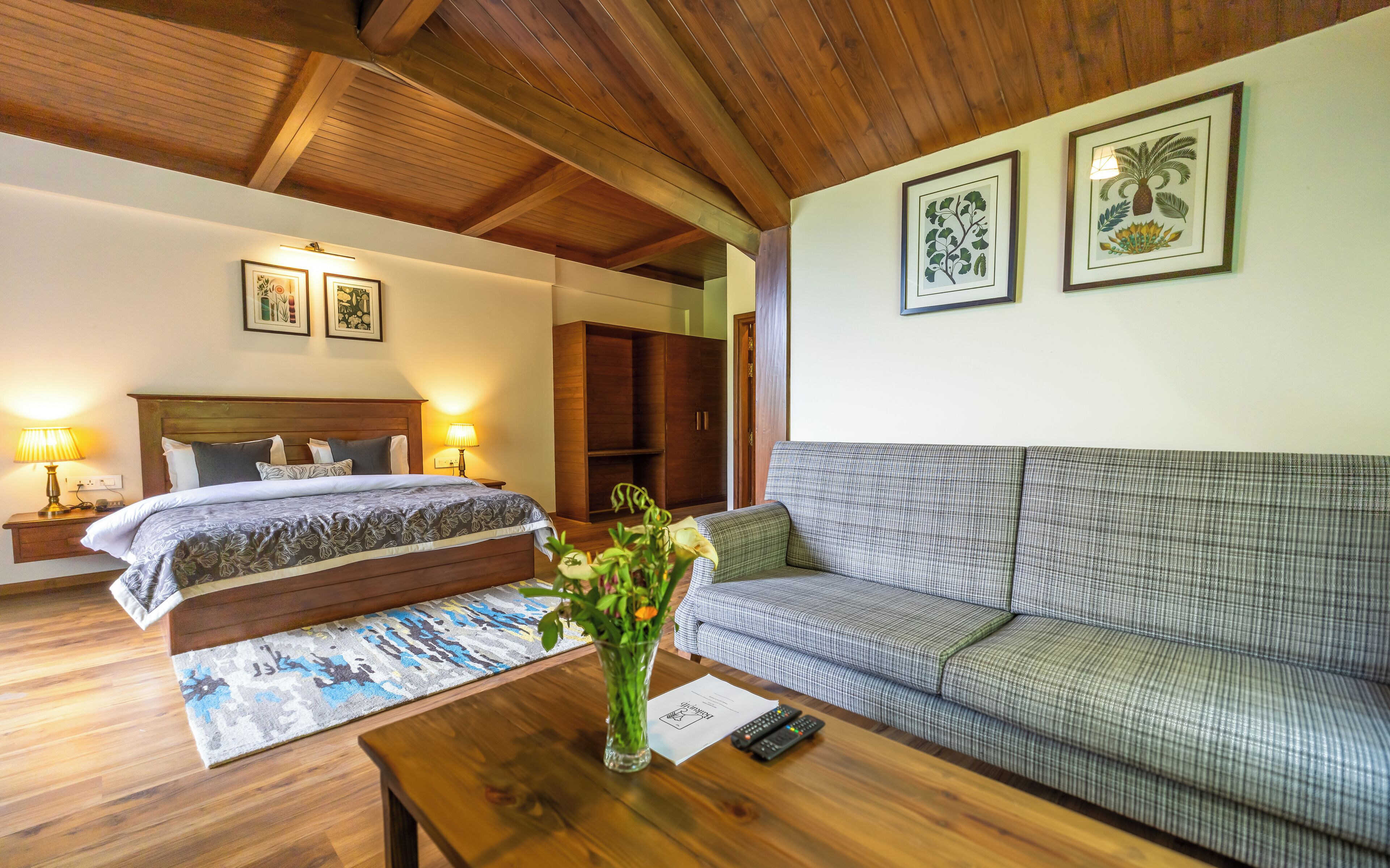 A hotel room with a bed, a sofa, a wooden coffee table, a vase with flowers, and framed botanical prints on the wall.