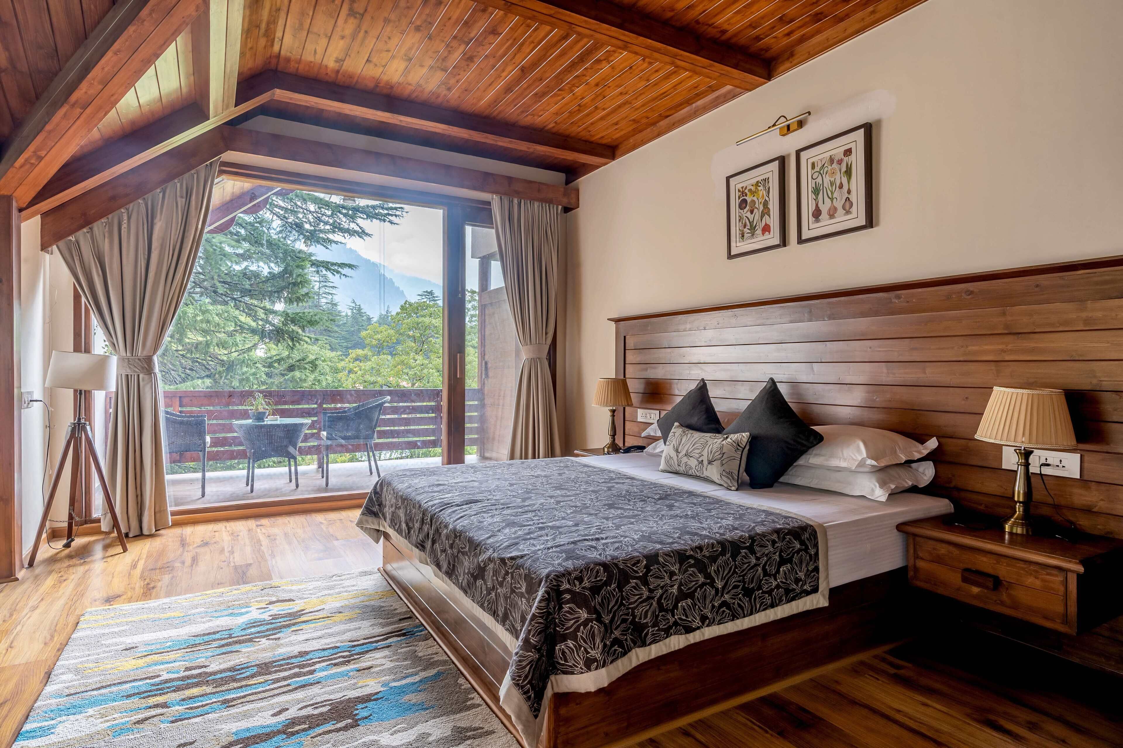 A bedroom with a wooden ceiling, a large bed with a patterned bedspread, a nightstand with a lamp, and a view of greenery through the window.
