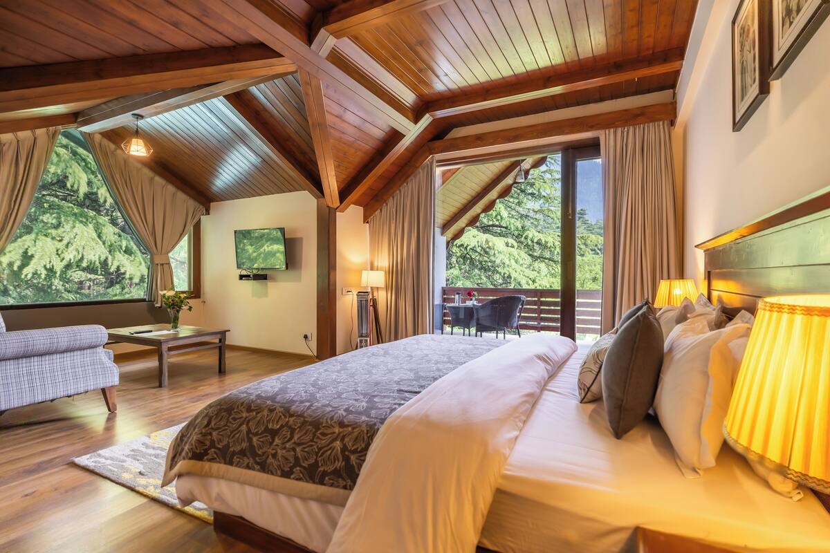 Un dormitorio amplio con una cama grande, un escritorio y una silla, un televisor montado en la pared y un balcón con vistas a la vegetación.
