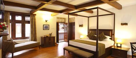 A four-poster bed with a bench, a small table, and a chair in a room with wooden floors and a large window.