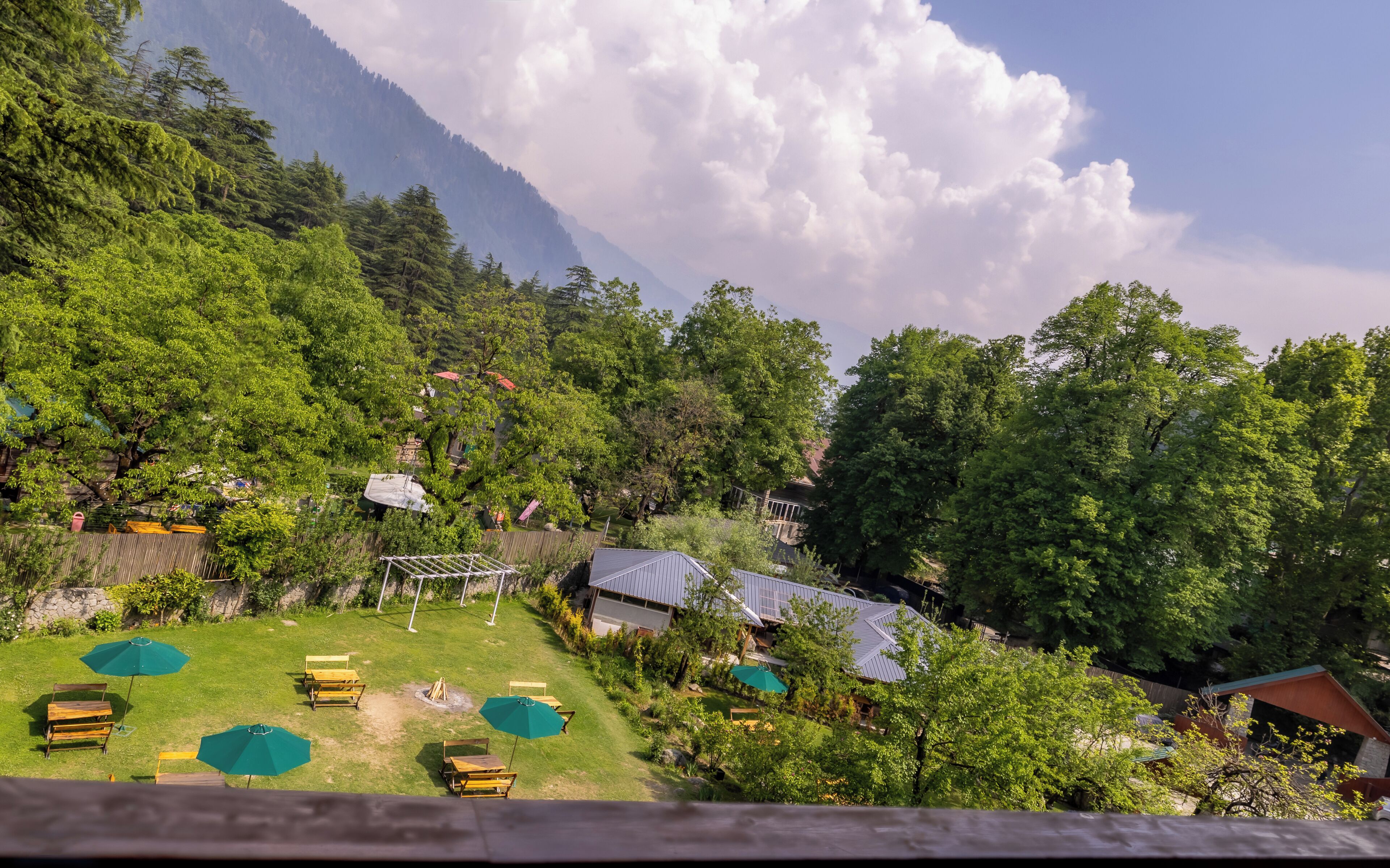 A lush green landscape with a mountain in the background, a grassy area with wooden benches and umbrellas, and a building with a metal roof.