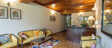 Uma sala de estar aconchegante com teto de madeira, um sofá amarelo, uma mesa de centro e uma pequena mesinha de cabeceira com abajur.