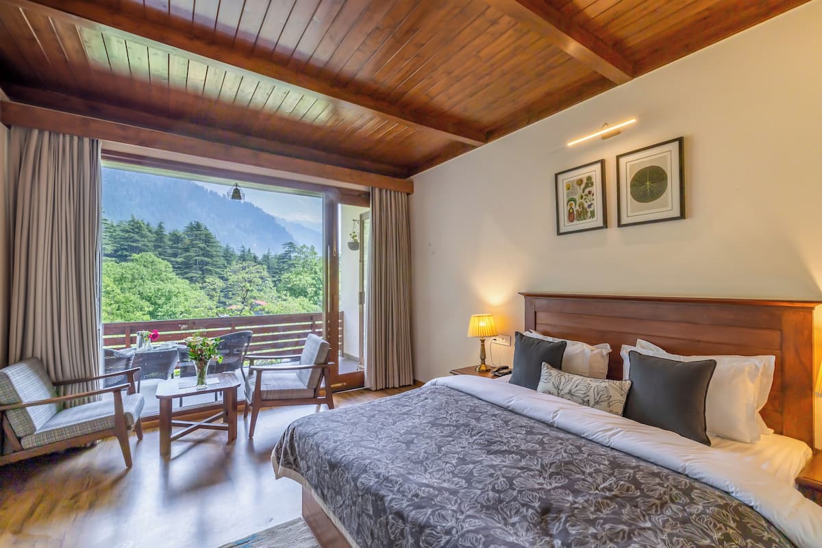 Habitación de hotel con una cama grande, un balcón con vistas a las montañas, una zona de comedor con sillas y una mesa, y una mesita de noche con una lámpara.