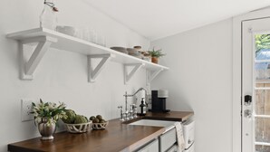 A kitchen with white cabinets, a wooden countertop, and a built-in sink. There are shelves above the countertop holding dishes and a potted plant. - Charming Downtown Country Cottage (Chico)