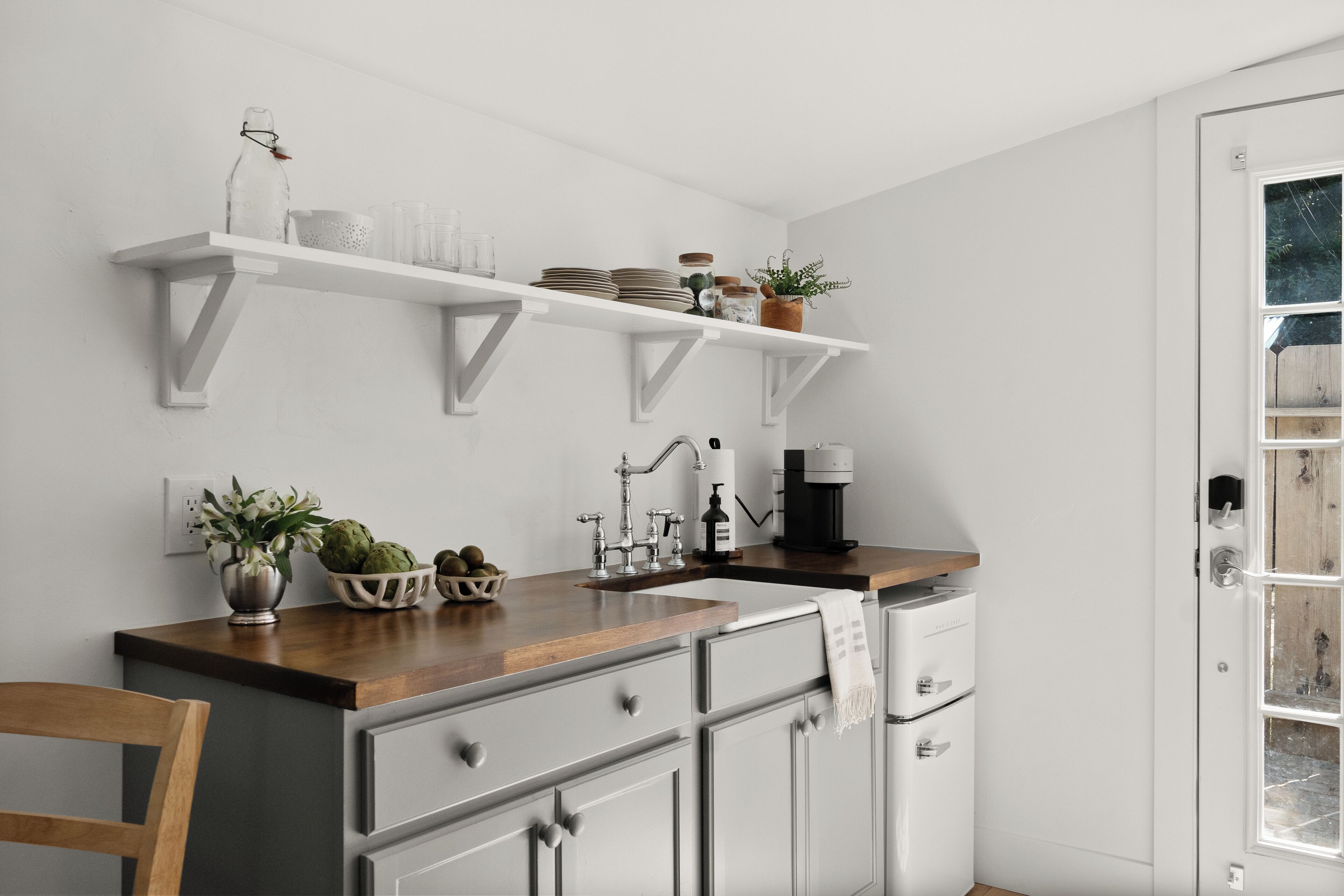 A kitchen with white cabinets, a wooden countertop, and a coffee maker. There are shelves above the sink holding plates and jars.