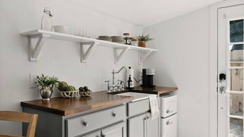 A kitchen with white cabinets, a wooden countertop, and a coffee maker. There are shelves above the sink holding plates and jars.