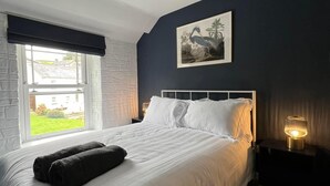 Un lit bien fait, avec du linge de lit blanc et une couverture sombre, une lampe de chevet, un tableau encadré au mur et une fenêtre donnant sur un paysage verdoyant.