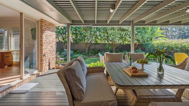 Un patio techado con una mesa de madera y mobiliario de mimbre, una pared de ladrillo y vistas a un jardín.