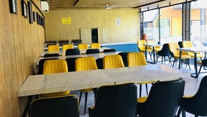 A cafeteria with tables and chairs, a wooden wall with framed pictures, and a blue wall with a window. - Eden Rock River Resort (Rishikesh)