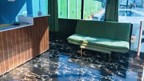 A reception area with a check-in/out desk, a green sofa, and a window with a view of greenery and a fence. - Eden Rock River Resort (Rishikesh)