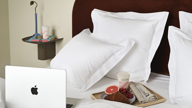 Un lit bien fait, avec du linge de lit blanc, un ordinateur portable, une tasse de café et des aliments pour le petit-déjeuner sur la table de chevet.