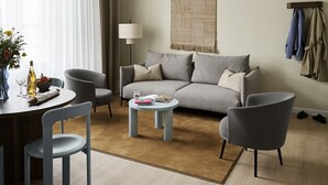 Un salon moderne comprenant un canapé, deux fauteuils, une table basse et une table à manger avec des chaises. On y trouve également une tapisserie murale et un porte-manteau avec des vêtements.