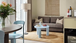 Ein modernes Wohnzimmer mit einer Couch, einem runden Couchtisch, einem Esstisch mit Stühlen, einer Lampe und einer Küchenzeile mit Geräten.