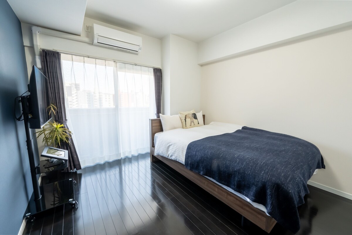 A modern hotel room with a large bed, a flat-screen TV, a small table with a plant, and a window with curtains.