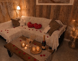A cozy room with a sectional sofa, a wooden coffee table, candles, and a bottle of champagne. - Gîte Maison de Campagne au nid D'ange Avec Jacuzzi (Saint-Michel-l'Observatoire)